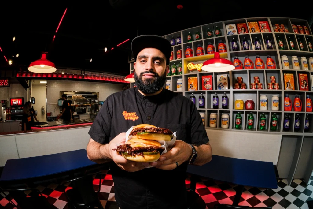 Abbas Dhanani holding two burgers inside the Katy restaurant, with the bodega-style interior visible behind him.