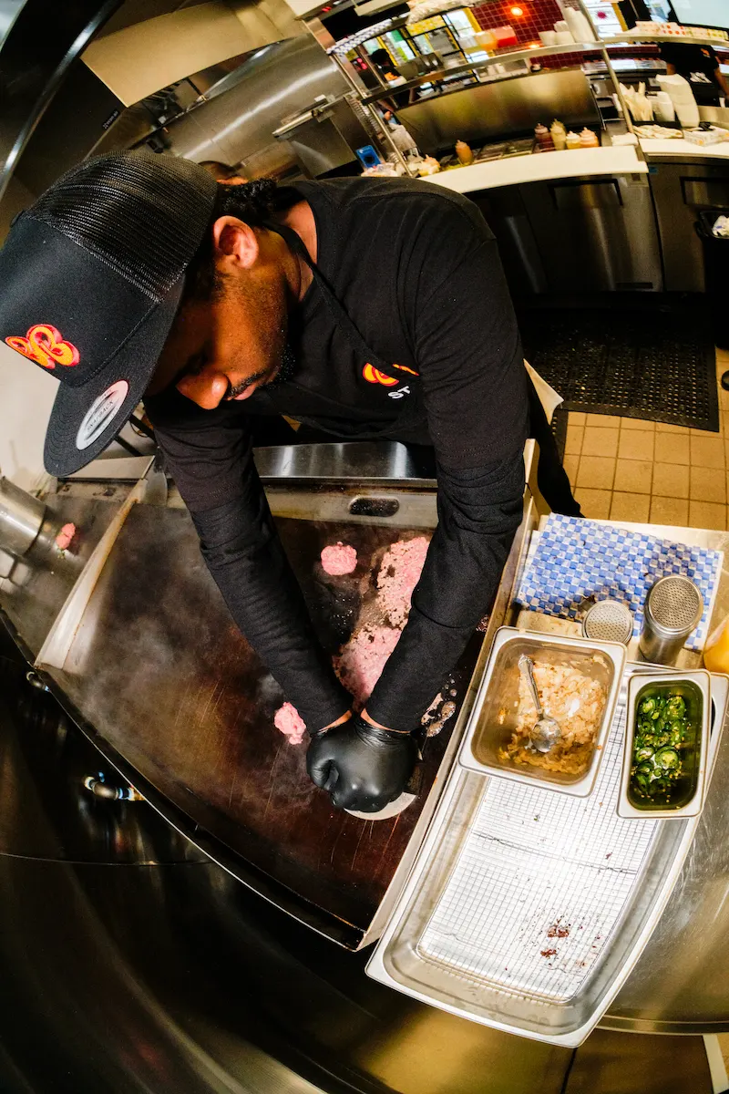 Burger Bodega staff member preparing smash burgers on a flat-top grill inside the kitchen.