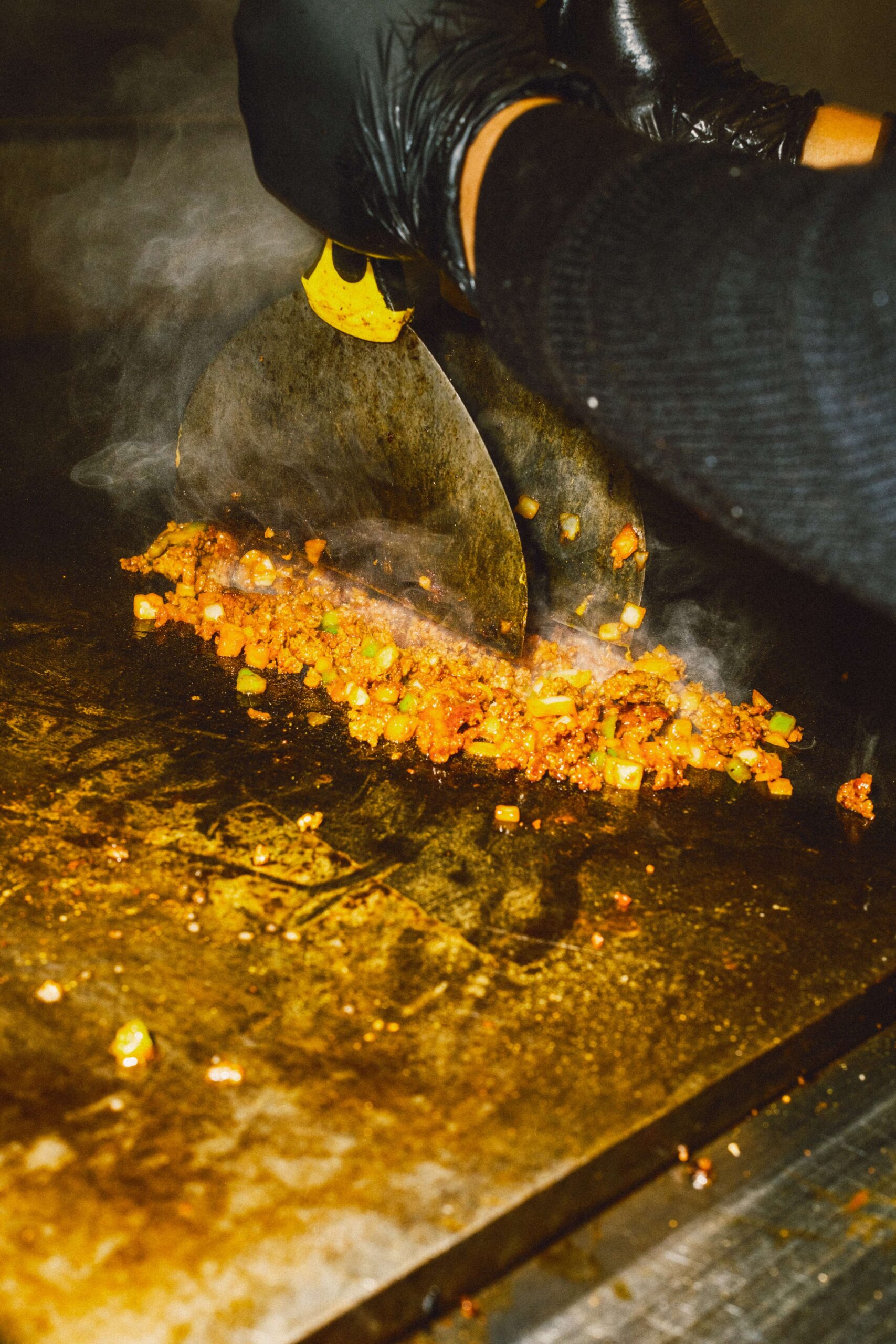 Burger patty mixture being cooked and chopped on a hot griddle with a metal spatula.